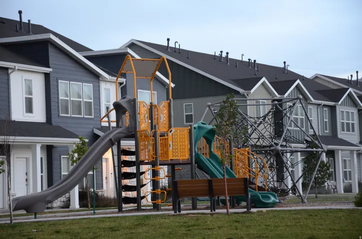 Community playground featuring a slide, climbing structure, and bench seating, set against a backdrop of modern townhomes with dark roofing and varied siding