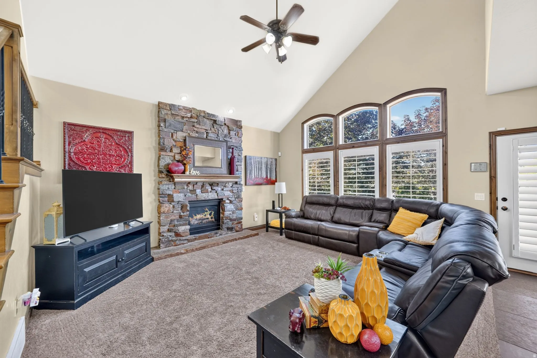 Vaulted ceiling living room featuring a floor-to-ceiling stone-clad fireplace, arched windows with plantation shutters, a ceiling fan, and carpet flooring