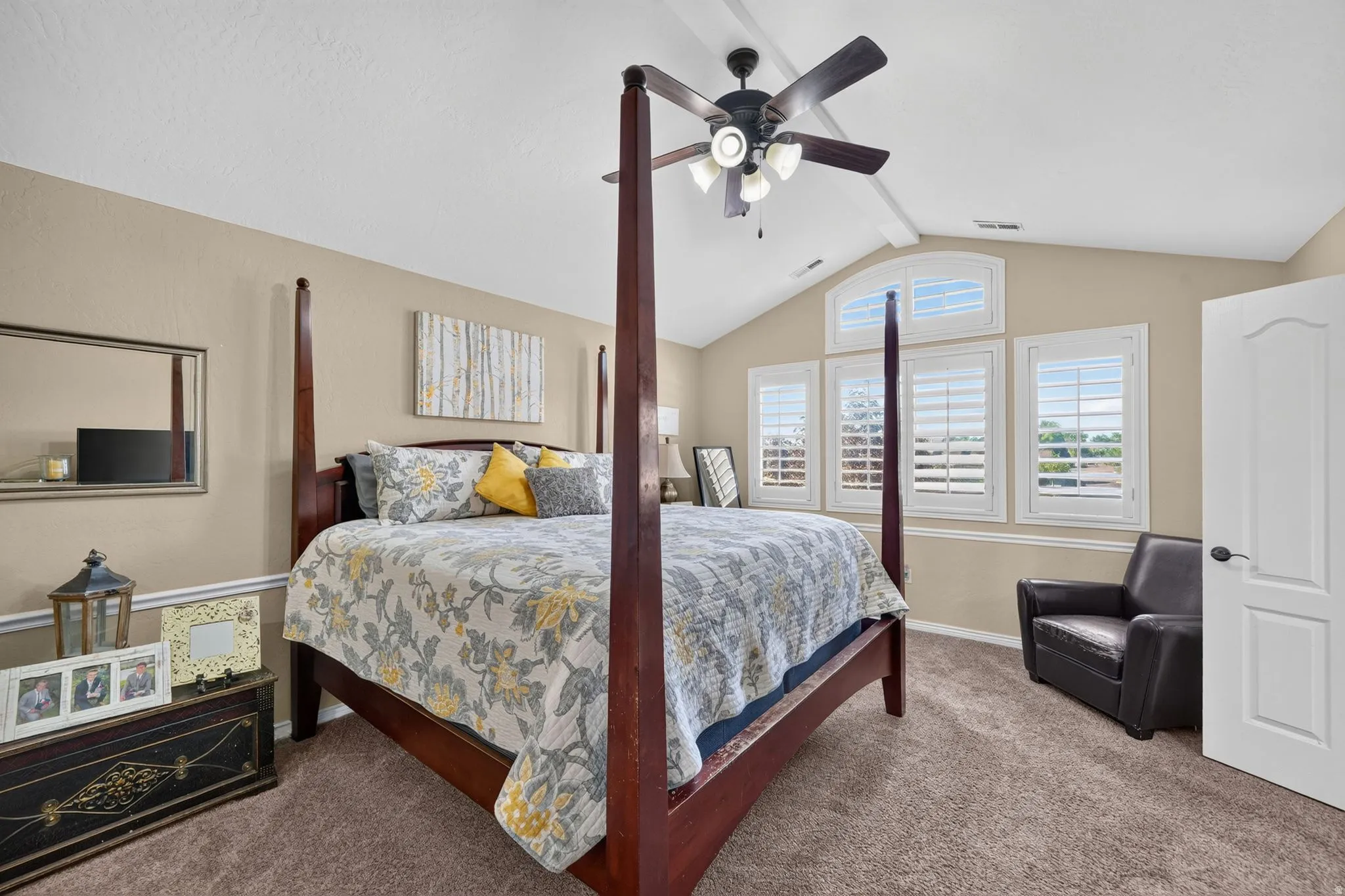 Primary bedroom with vaulted ceiling featuring a ceiling fan with integrated lighting, multiple windows with plantation shutters, warm neutral wall tones, and plush carpeting