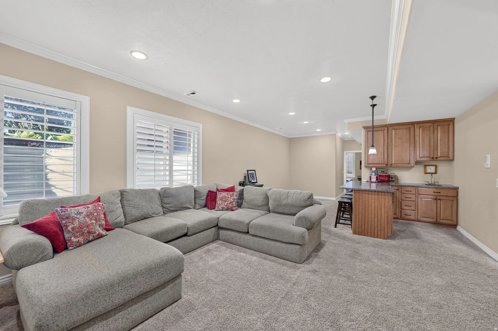 Basement living room with recessed lighting illuminates this carpeted entertainment space, featuring two windows with plantation shutters, a built-in wet bar with wood cabinetry, and a countertop with an integrated sink