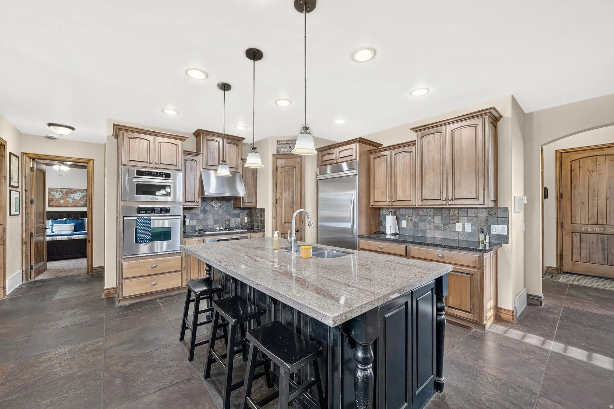 Gourmet kitchen featuring a large central island with seating, stainless steel appliances, wood cabinetry, stone countertops, and a tiled backsplash