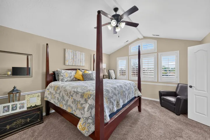 Primary bedroom with vaulted ceiling featuring a ceiling fan with integrated lighting, multiple windows with plantation shutters, warm neutral wall tones, and plush carpeting