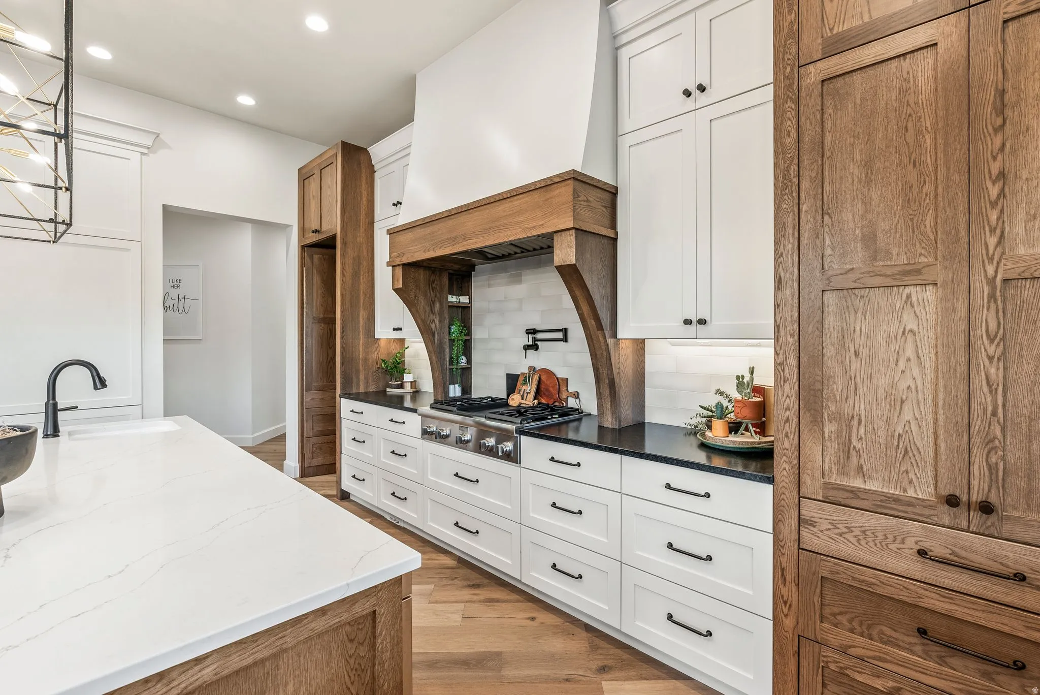 Spacious kitchen featuring an island with a white countertop and undermount sink, a gas range with a pot filler faucet, a custom wood hood, white shaker cabinetry, and wood-finish flooring