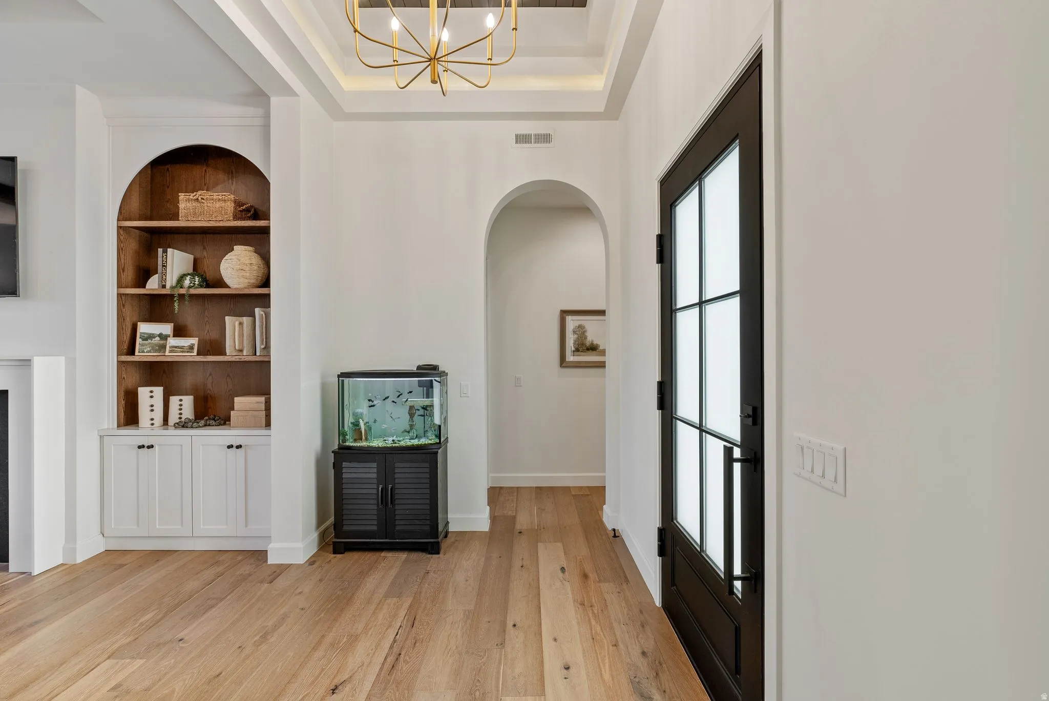 Entryway featuring a dark-framed full-light door with frosted panels, light wood-finish flooring, a coffered ceiling with a gold-tone chandelier, and an arched built-in bookshelf with lower cabinetry
