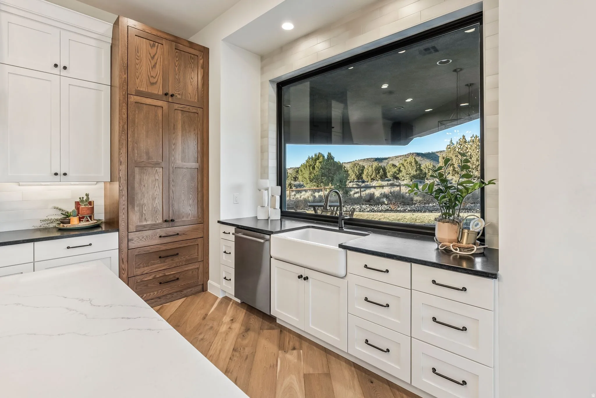 Kitchen featuring a wide picture window, farmhouse sink, wood-finish flooring, white cabinetry with black hardware, and a built-in pantry with wood-finish doors