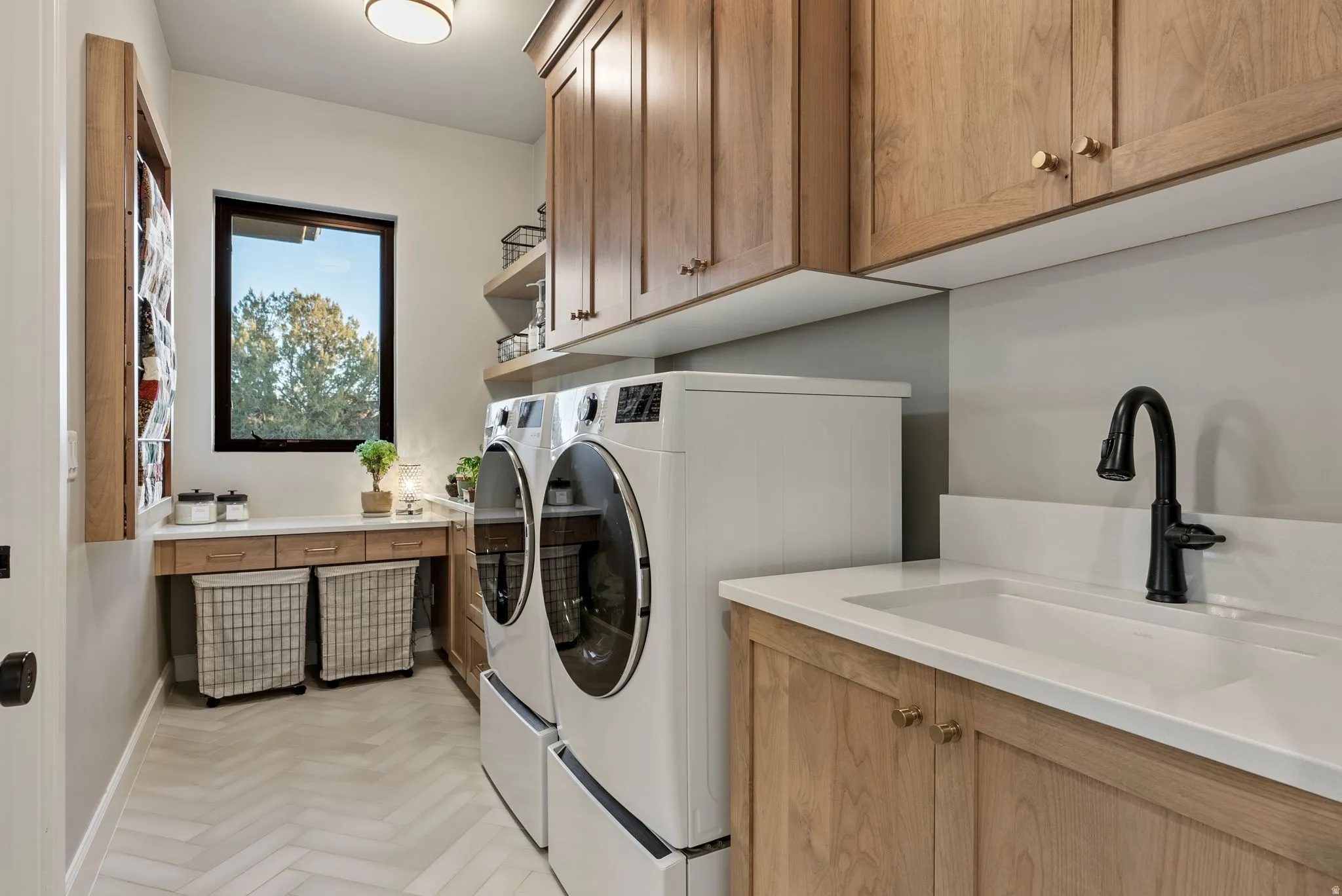 Functional laundry room featuring herringbone pattern flooring, natural wood cabinetry, white countertops, built-in sink with matte black faucet, and a large window