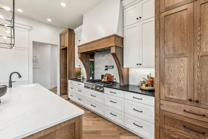 Spacious kitchen featuring an island with a white countertop and undermount sink, a gas range with a pot filler faucet, a custom wood hood, white shaker cabinetry, and wood-finish flooring