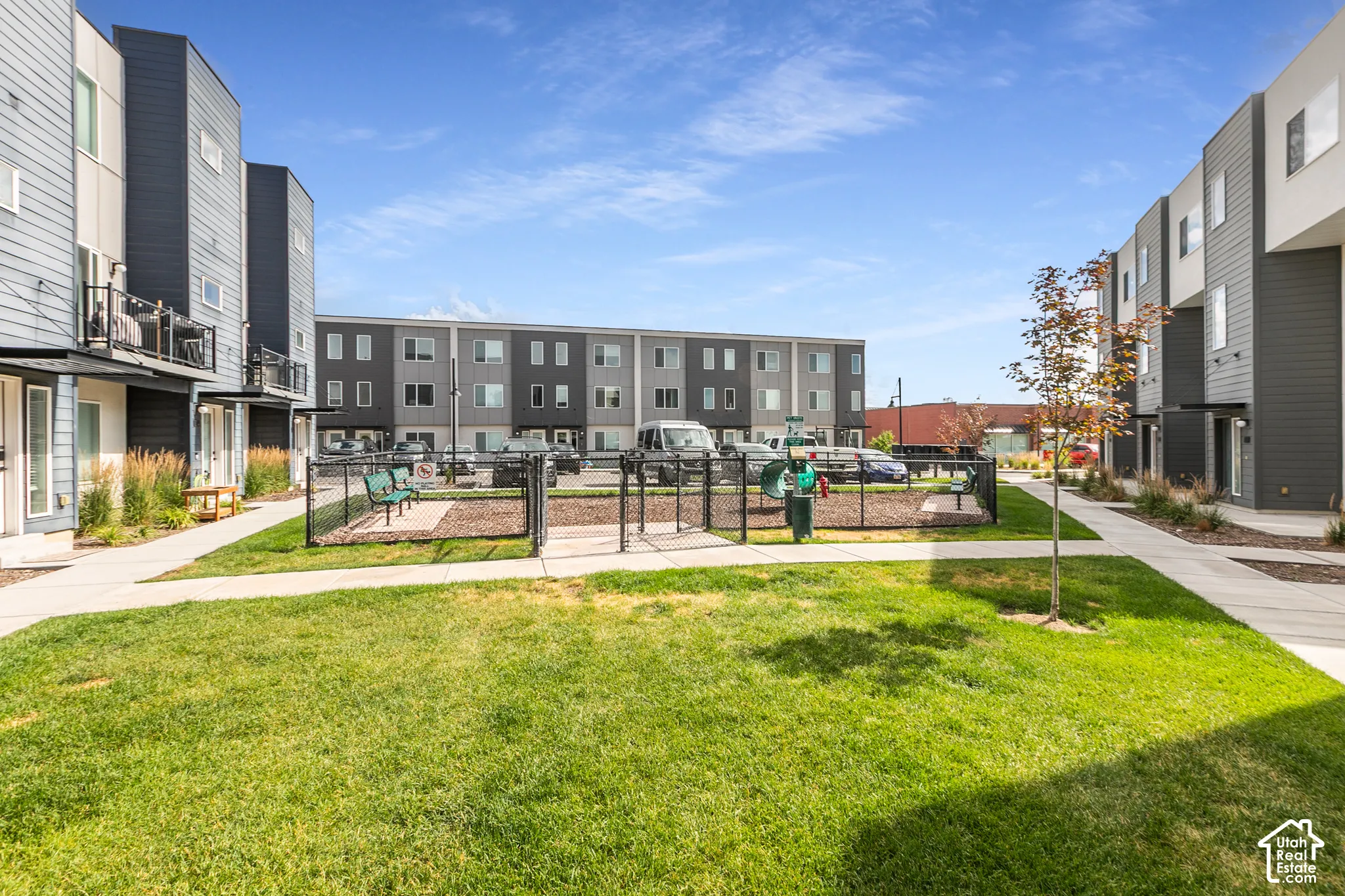 Community green space featuring a manicured lawn, paved walkways, and a fenced pet area with benches