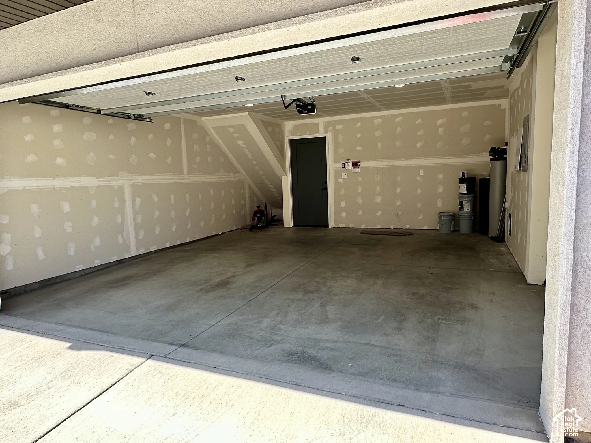 Concrete garage flooring, unfinished drywall, interior door, overhead garage door mechanism, and recessed ceiling lighting