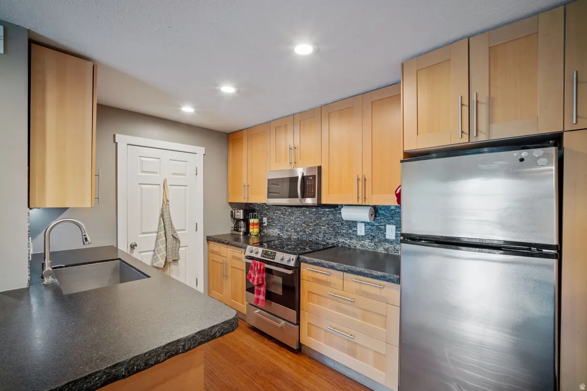 Kitchen featuring light wood cabinetry, dark countertops, stainless steel appliances, a mosaic tile backsplash, and recessed lighting