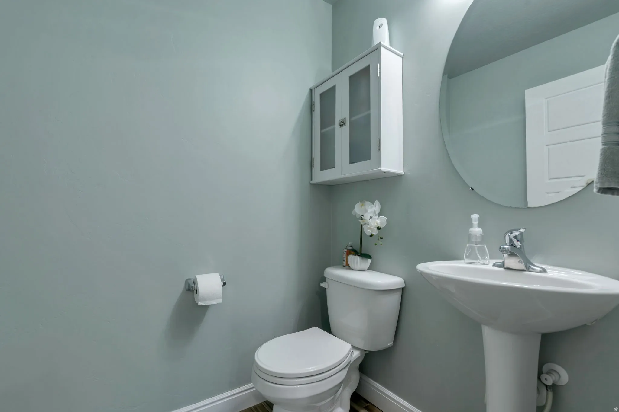Pedestal sink with chrome faucet, circular wall mirror, and wall-mounted storage cabinet with frosted glass panels