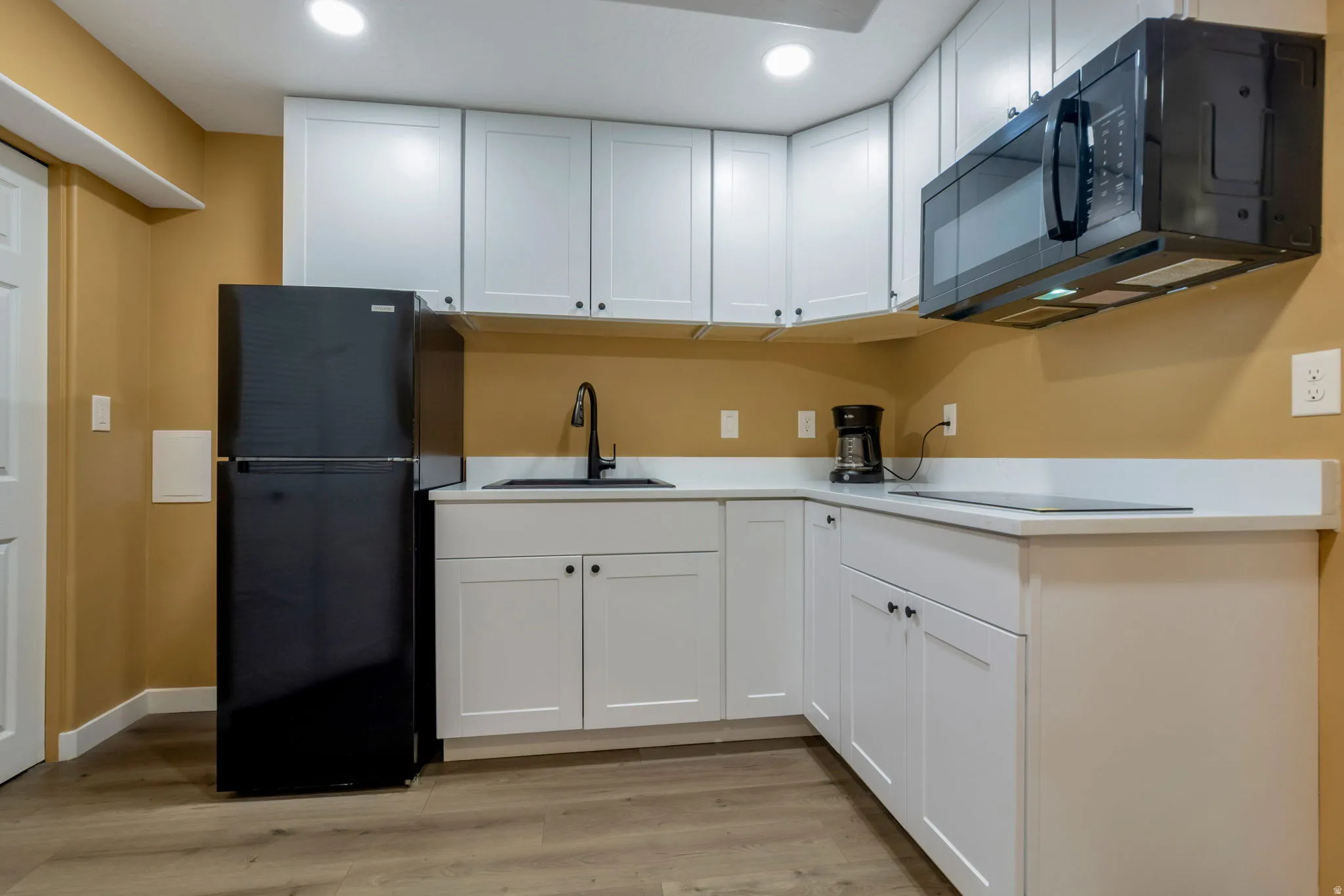 Compact kitchen featuring white shaker-style cabinetry, white countertops, a black top-freezer refrigerator, a black over-range microwave, and wood-finish flooring