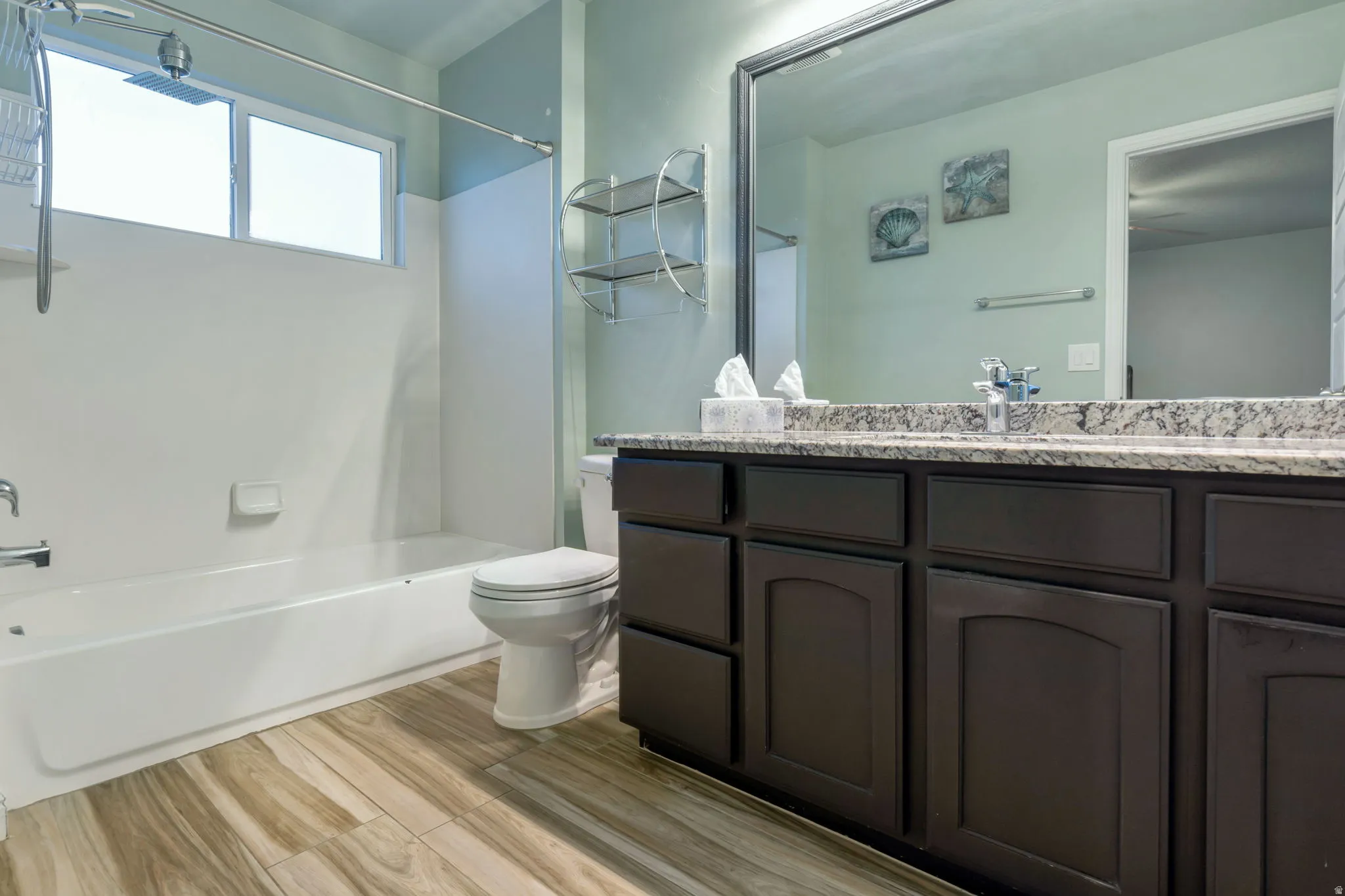 Bathroom featuring a dual vanity with a granite-style countertop, a built-in tub with shower, wood-finish flooring, and natural light from a horizontal window