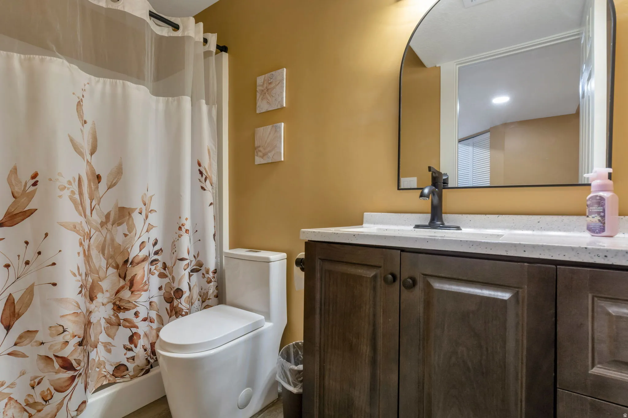 Bathroom featuring a wood-finish vanity with a light-toned countertop, an arched mirror, and a modern toilet