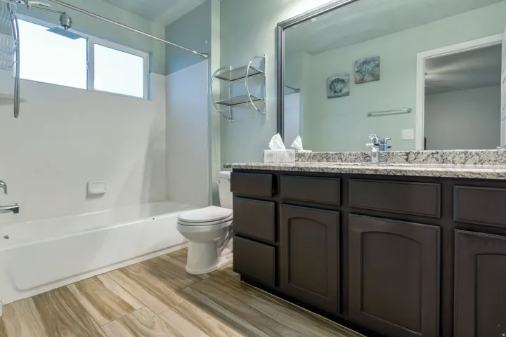 Bathroom featuring a dual vanity with a granite-style countertop, a built-in tub with shower, wood-finish flooring, and natural light from a horizontal window