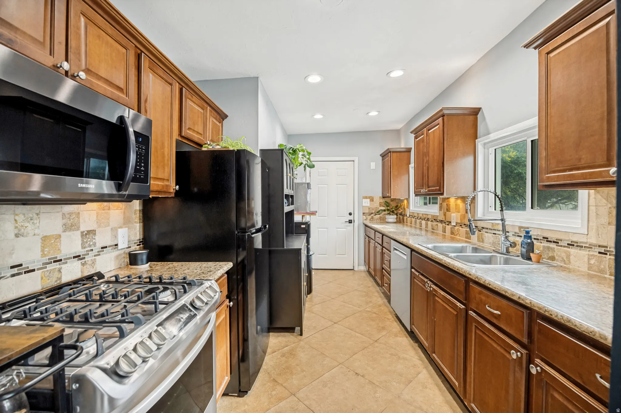 Galley kitchen featuring wood cabinetry, stainless steel range and microwave, tile countertops, and a mosaic tile backsplash