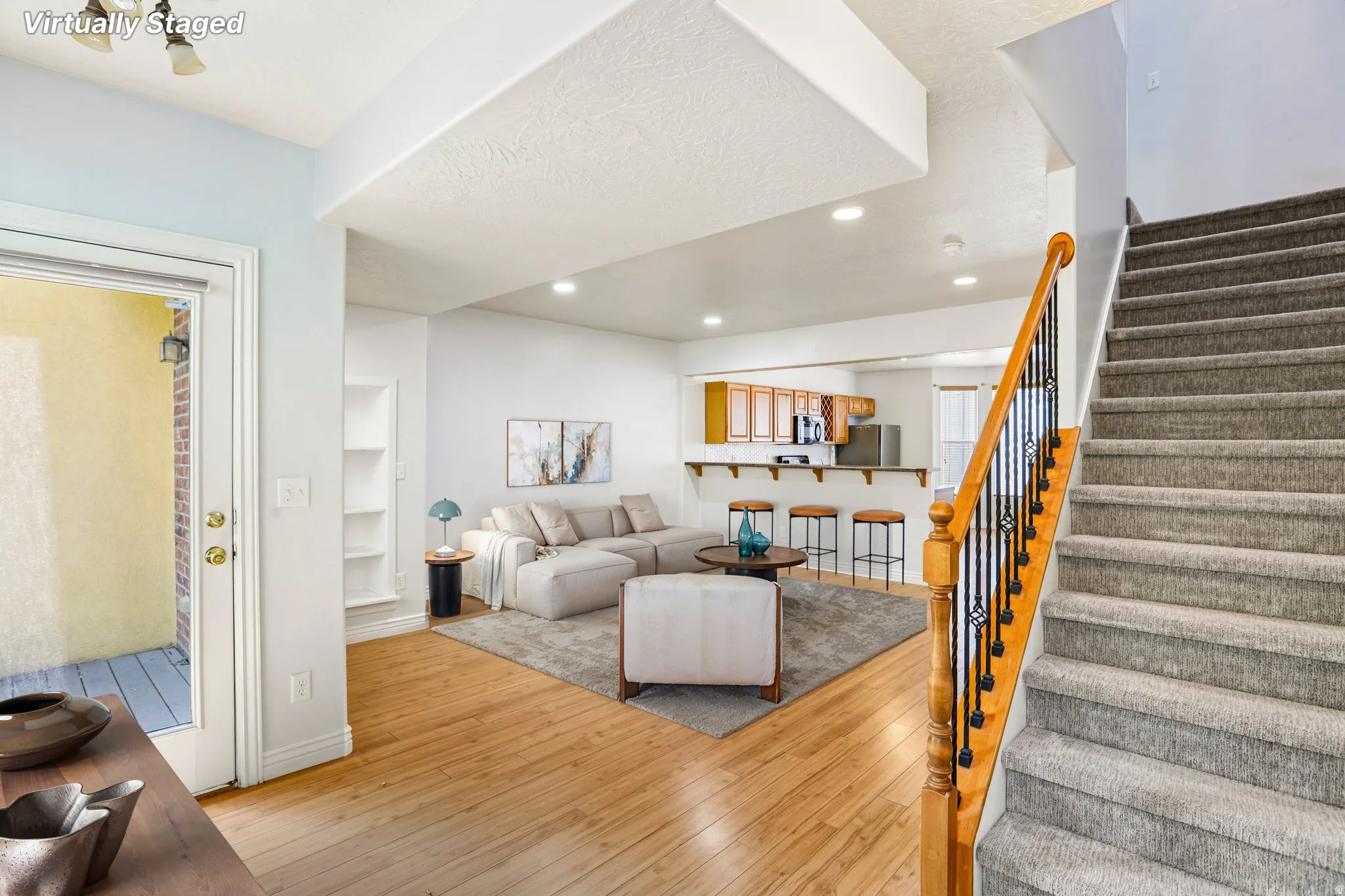 Open-concept living space featuring wood-finish flooring, a built-in shelving unit, recessed lighting, and a staircase with carpeted treads and an ornate metal and wood handrail