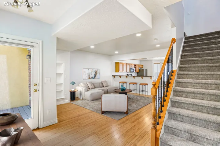 Open-concept living space featuring wood-finish flooring, a built-in shelving unit, recessed lighting, and a staircase with carpeted treads and an ornate metal and wood handrail