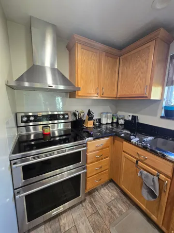 Stainless steel double oven range with matching range hood, light wood cabinetry, black countertops, and wood-finish flooring
