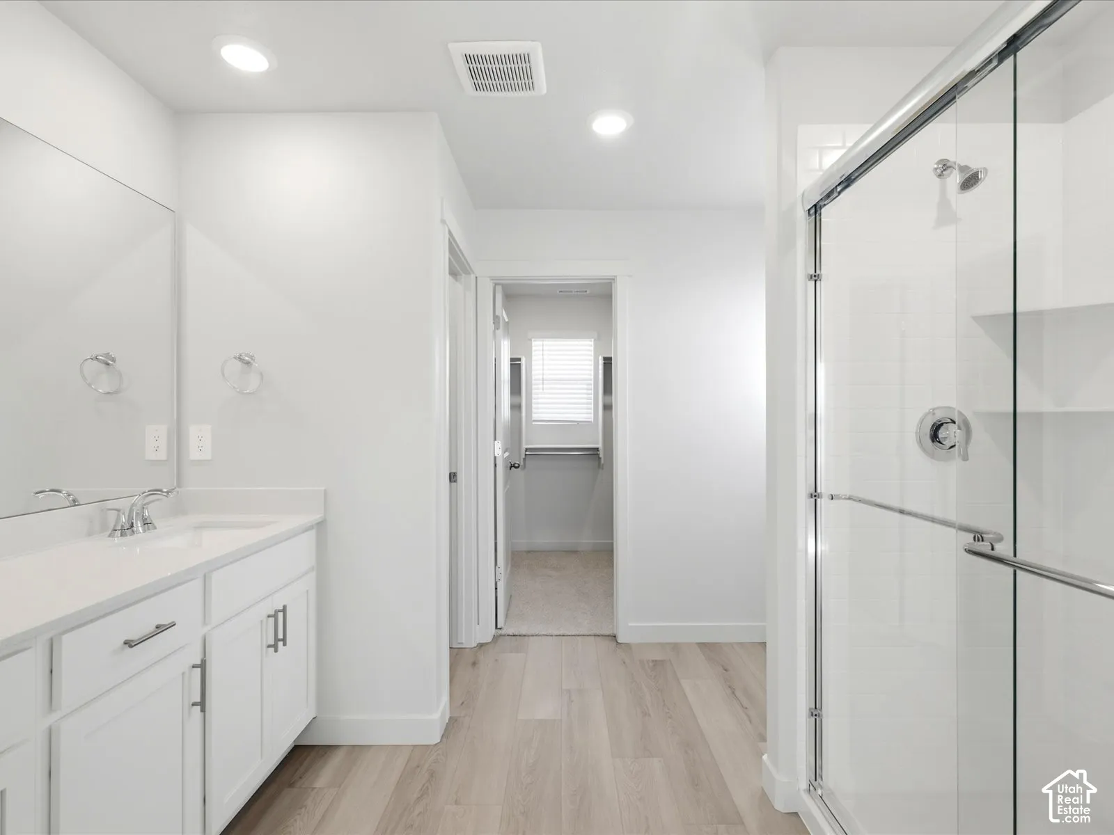 Bathroom featuring wood-finish flooring, white cabinetry with a light-toned countertop, a full-width mirror, recessed lighting, and a glass-enclosed shower