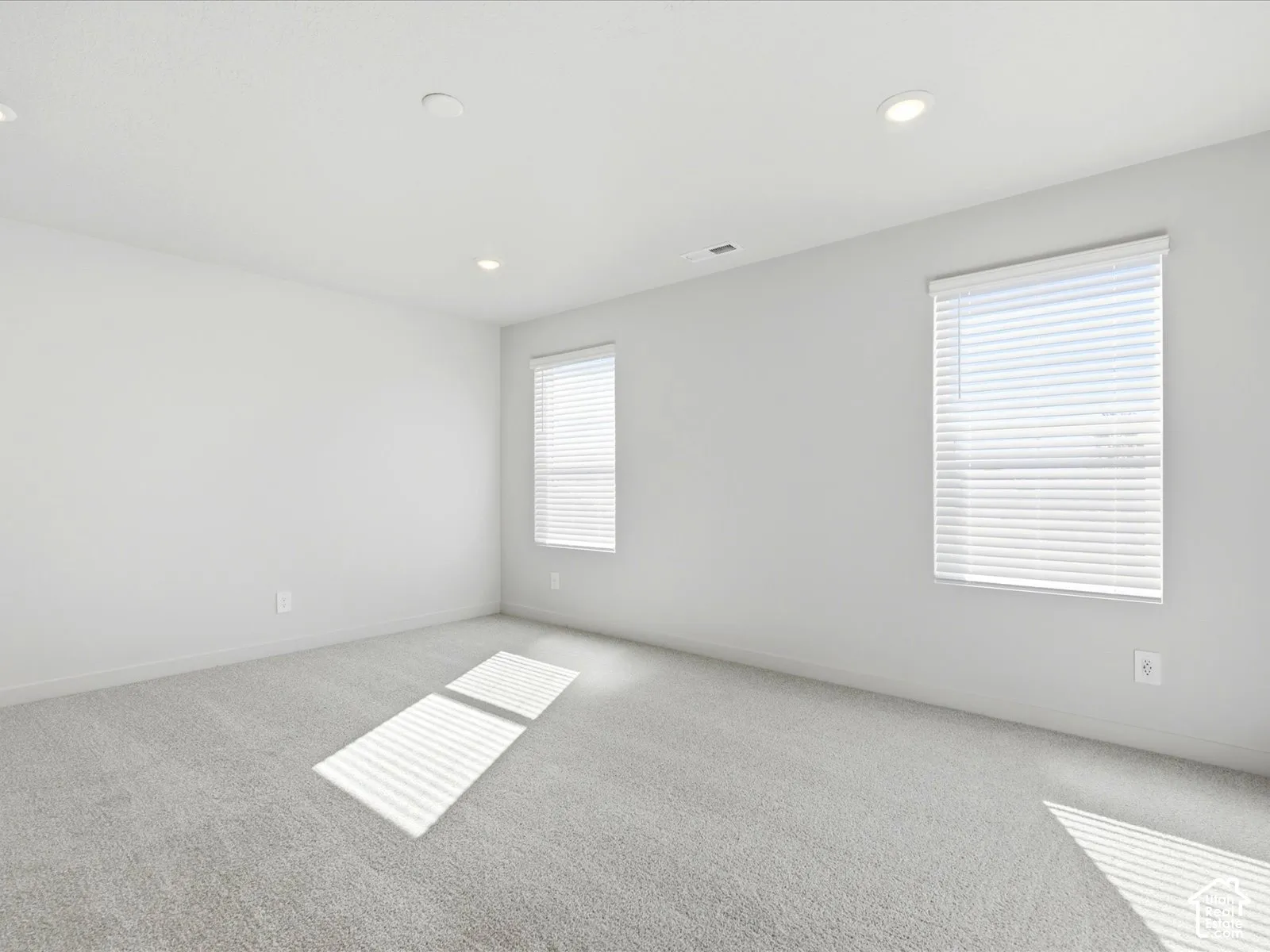 Neutral-toned interior space featuring light wall-to-wall carpeting, recessed ceiling lighting, and dual windows with horizontal blinds