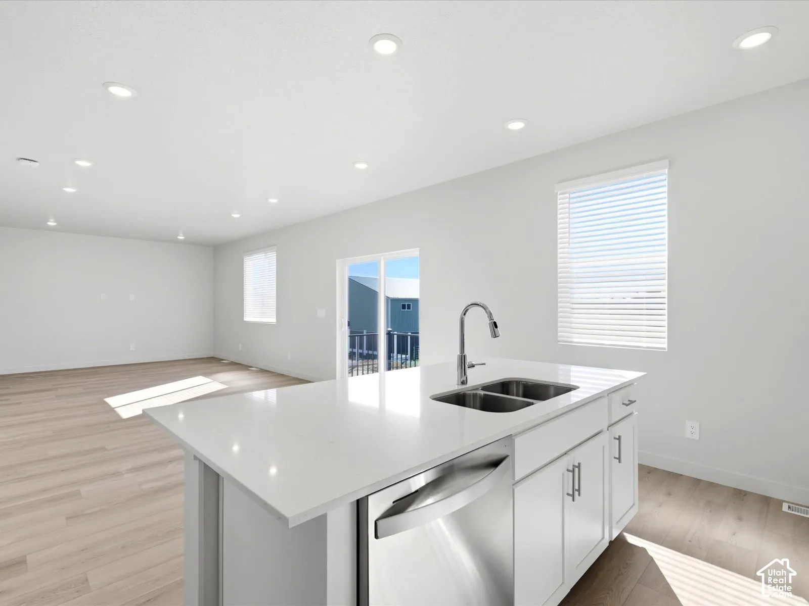 Kitchen island featuring a stone-composite countertop, dual-basin stainless steel sink, gooseneck faucet, integrated dishwasher, and white cabinetry, complemented by wood-finish flooring and recessed lighting