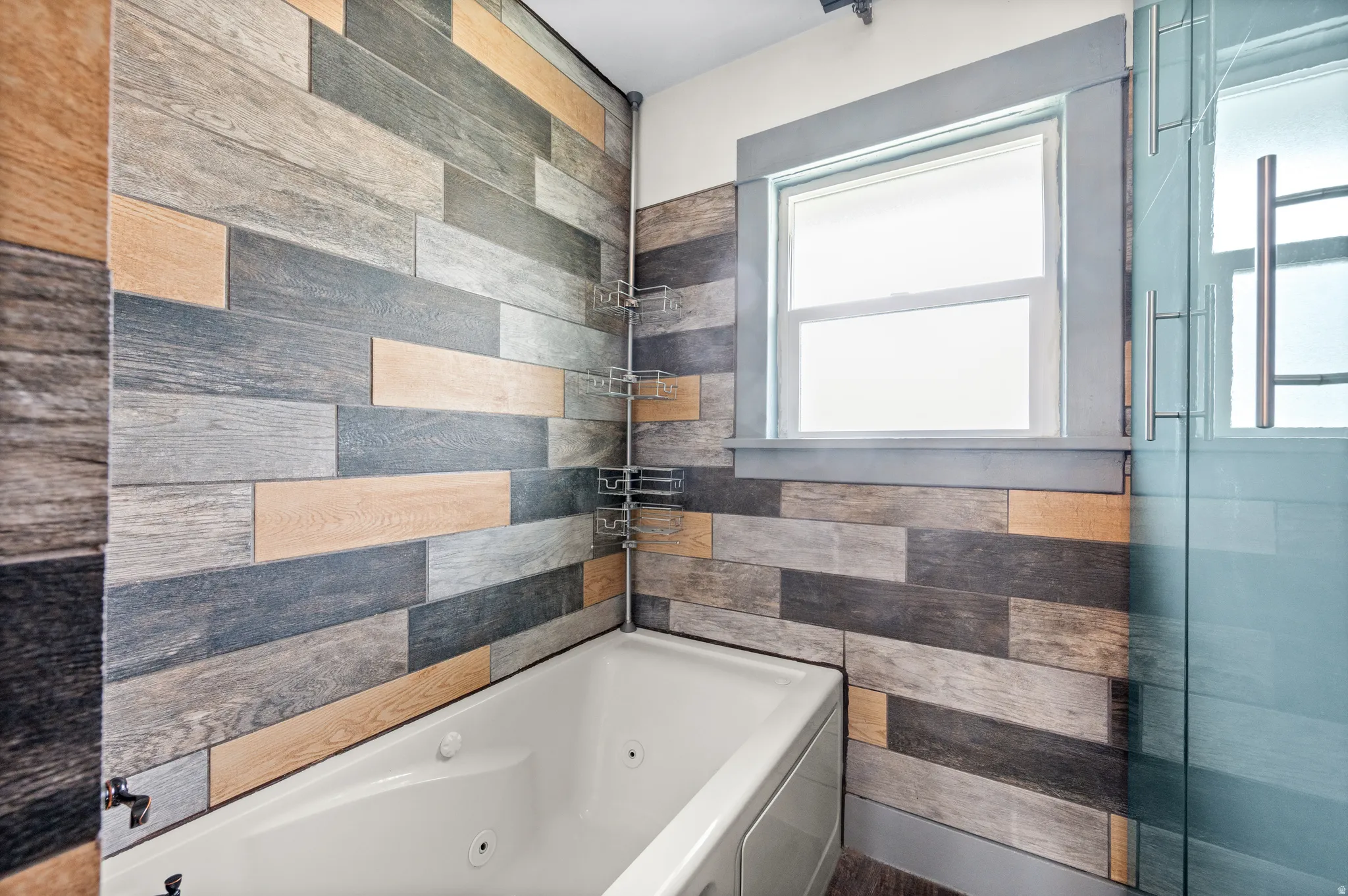 Soaking tub with integrated jets, surrounded by wood-look plank tile walls in varied tones, a framed window, and clear glass shower enclosure