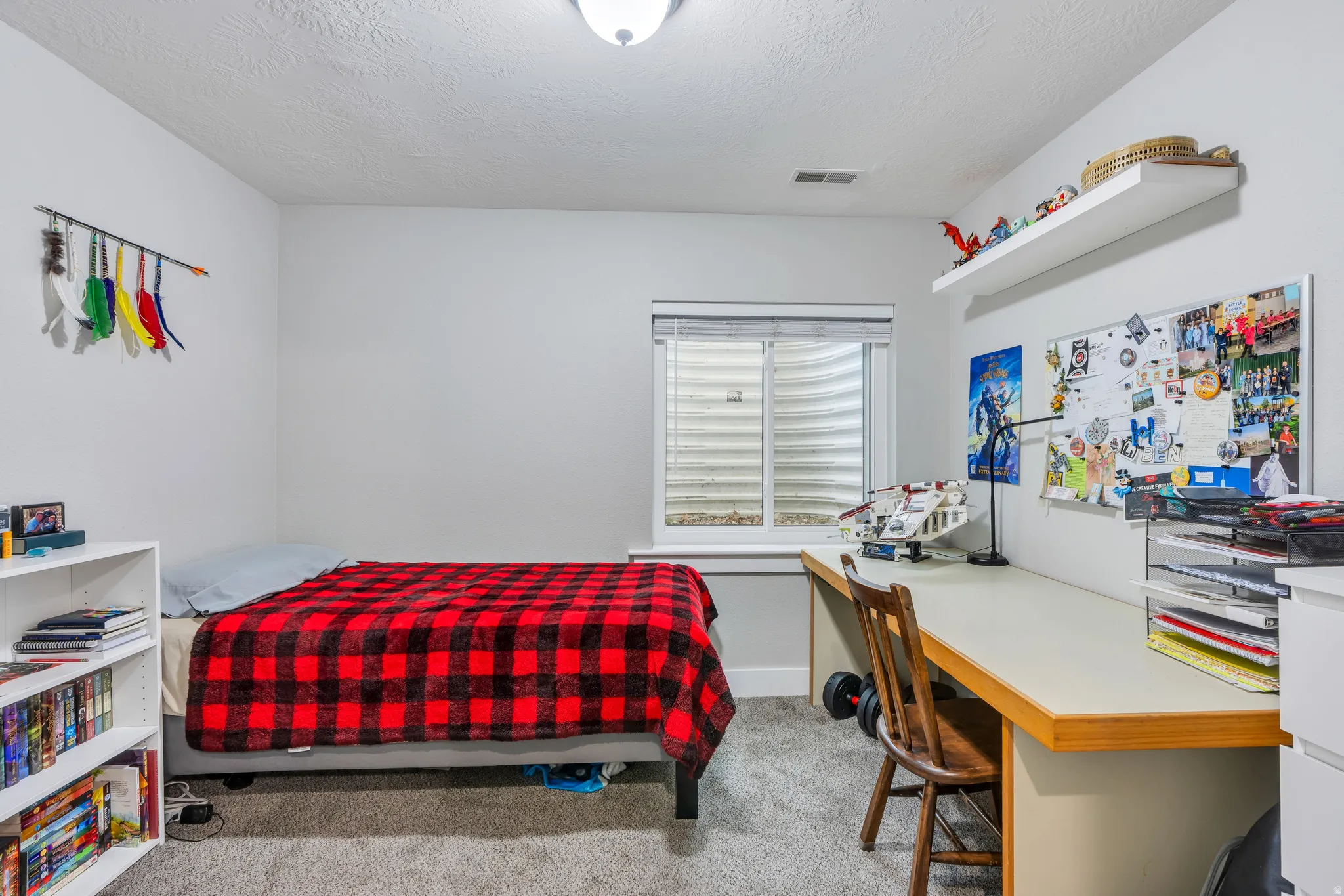 Room featuring light gray walls, plush gray carpeting, a built-in window with a horizontal blind, a built-in desk with a light-colored surface, and a wall-mounted floating shelf
