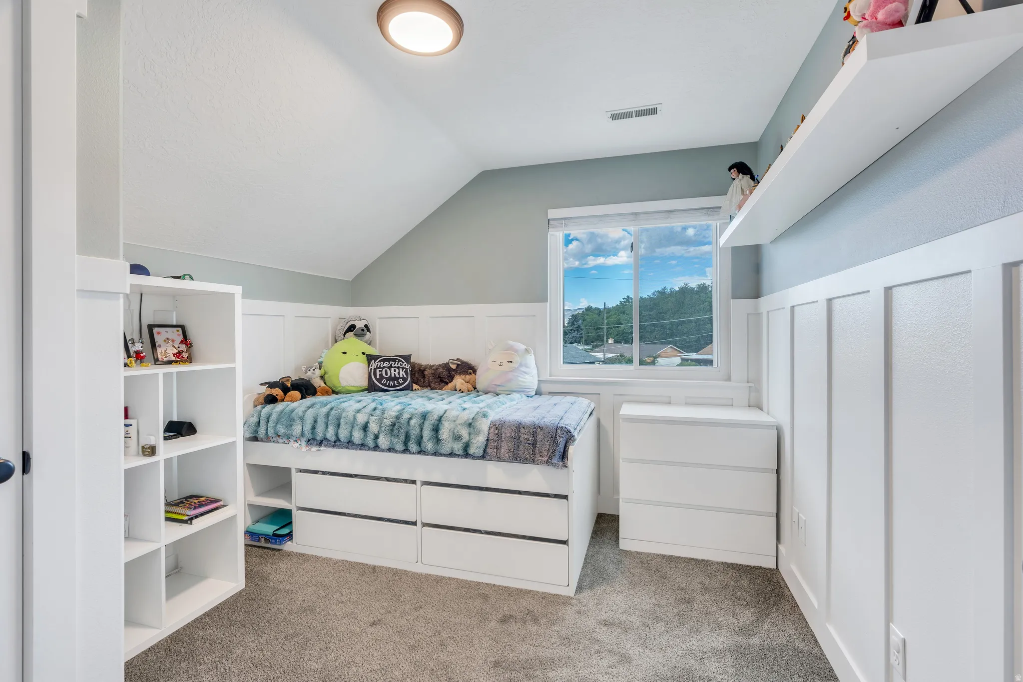 Angled ceiling bedroom featuring white wainscoting, an integrated window, a built-in shelving unit, a floating wall shelf, and a ceiling-mounted light fixture