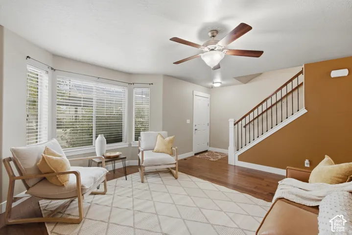 Bay window with horizontal blinds, wood-finish flooring, ceiling fan with light fixture, white trim, and a staircase with dark wood handrail and white balusters