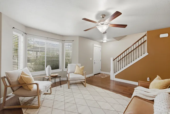 Bay window with horizontal blinds, wood-finish flooring, ceiling fan with light fixture, white trim, and a staircase with dark wood handrail and white balusters