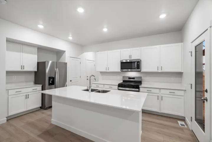 Bright kitchen featuring a central island with a double sink, white cabinetry with brushed nickel hardware, light-toned countertops, stainless steel appliances, and wood-finish flooring