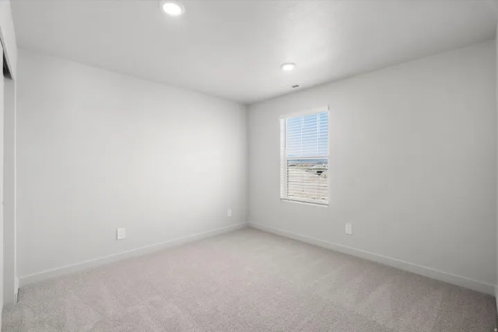 Bright secondary bedroom showcasing neutral carpeting, recessed ceiling lighting, and a single window framed with horizontal blinds allowing natural light throughout the space