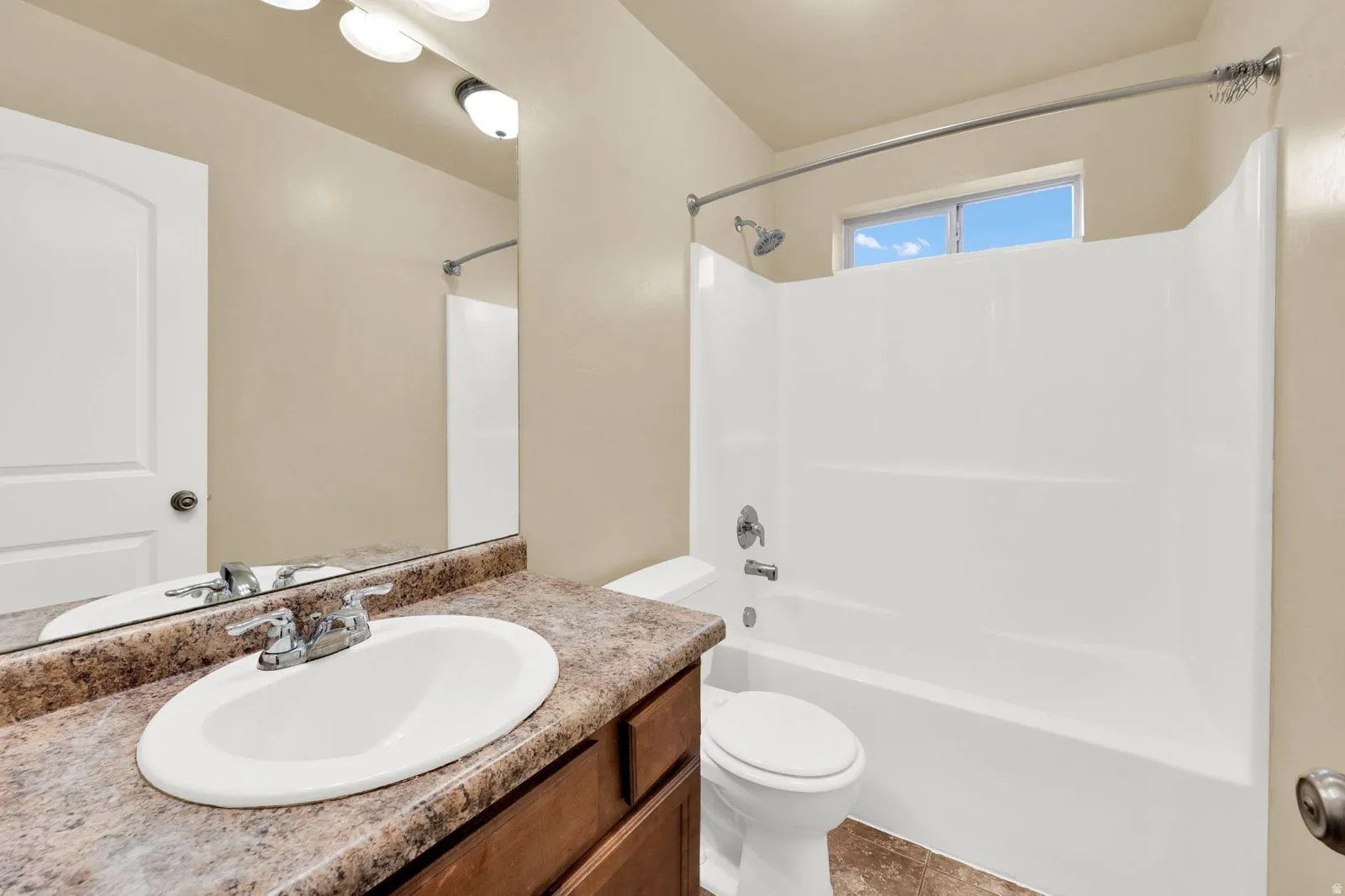 Bathroom featuring a wood-finish vanity with an oval sink and chrome faucet, a large vanity mirror, a combination tub and shower with white surround, and an upper window providing natural light