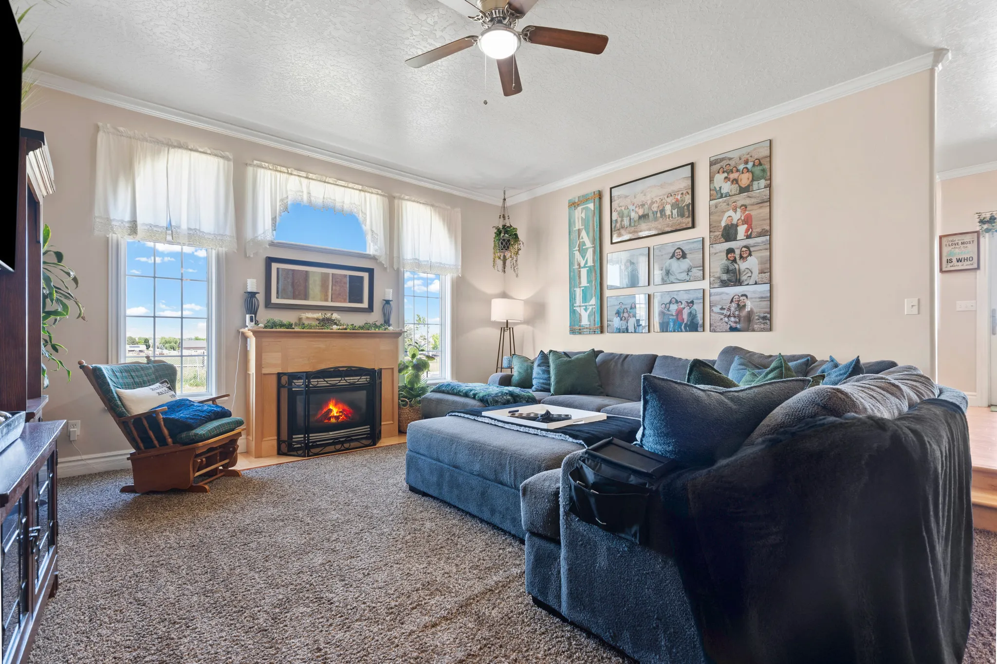 Living area featuring a fireplace with a wood mantel, multiple windows, a ceiling fan, and carpet flooring
