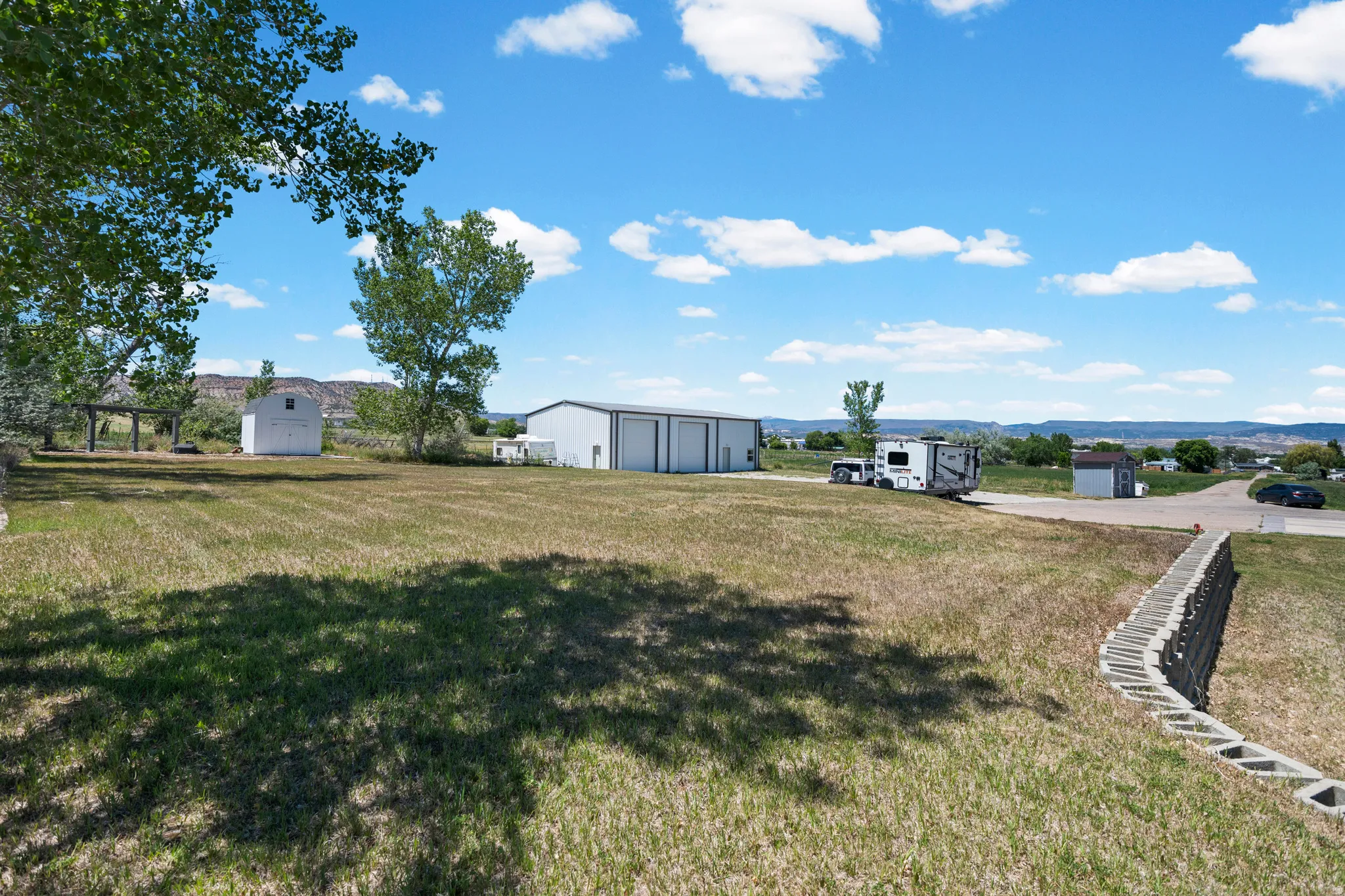 Expansive green space featuring a large metal-sided outbuilding with multiple bay doors, a white storage shed, and a retaining wall