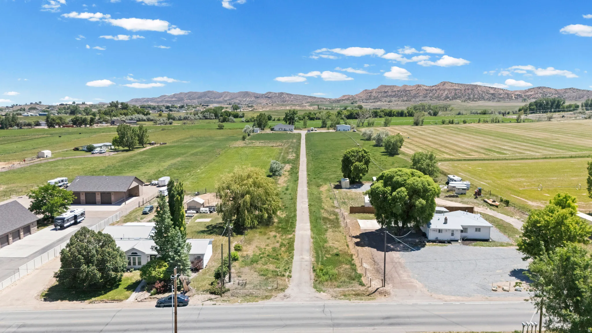 Expansive rural property featuring an elongated driveway, multiple structures, and a backdrop of verdant fields and distant mountains