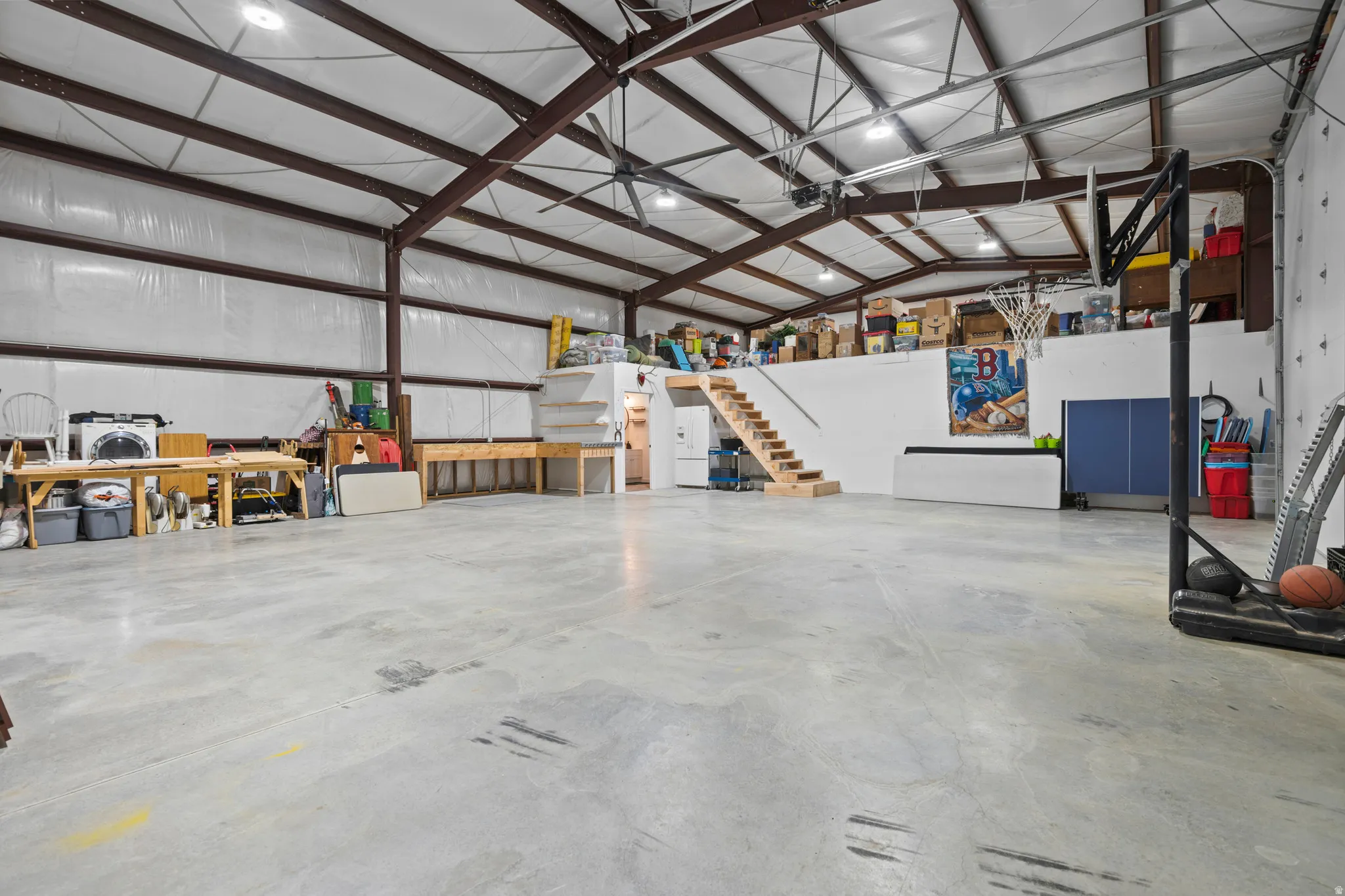 Spacious concrete-floored interior featuring high ceilings with exposed steel beams, industrial lighting, and a prominent mezzanine level with wood stairs