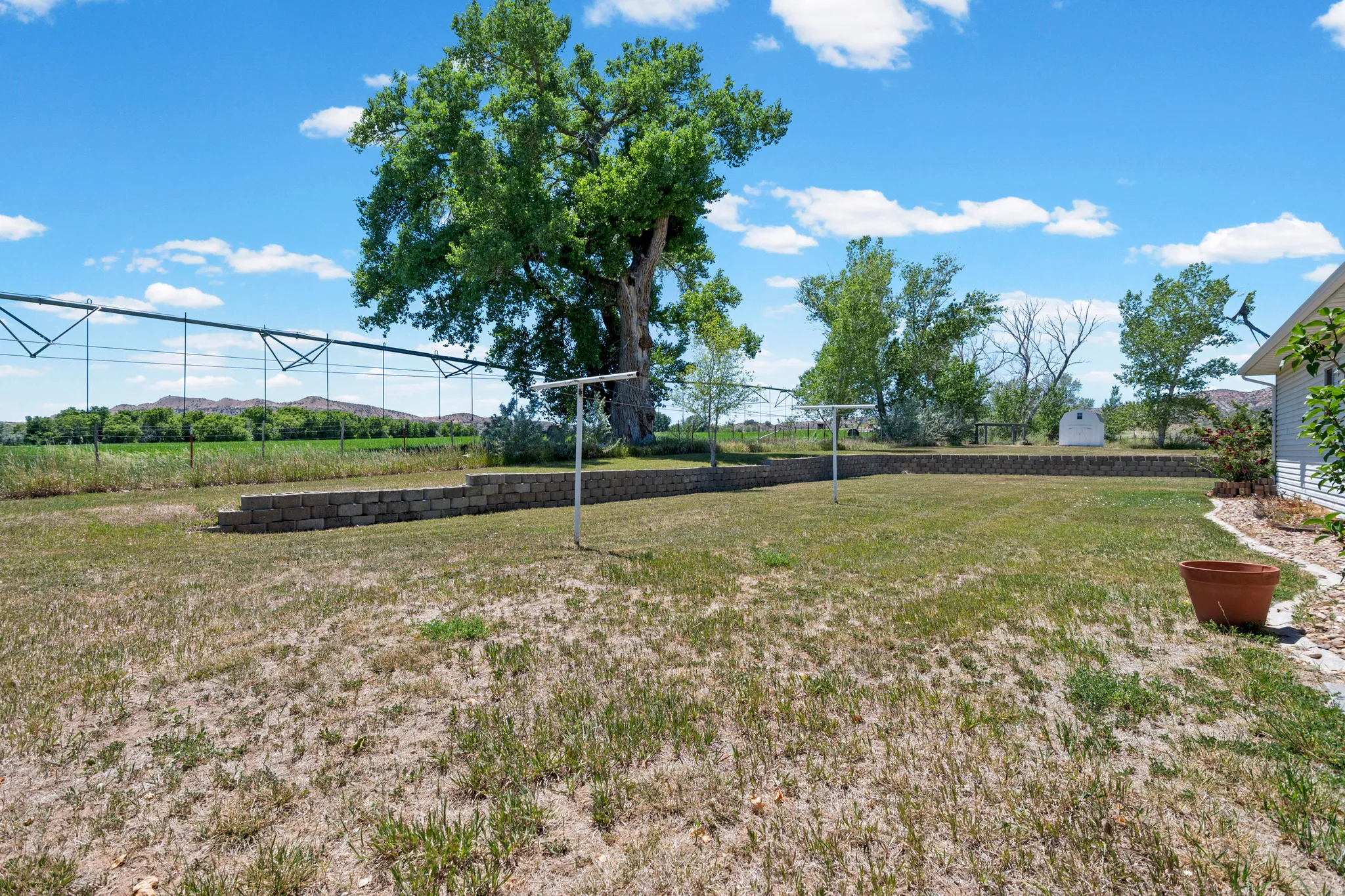 Expansive backyard featuring a retaining wall, mature trees, and a distant field with irrigation equipment