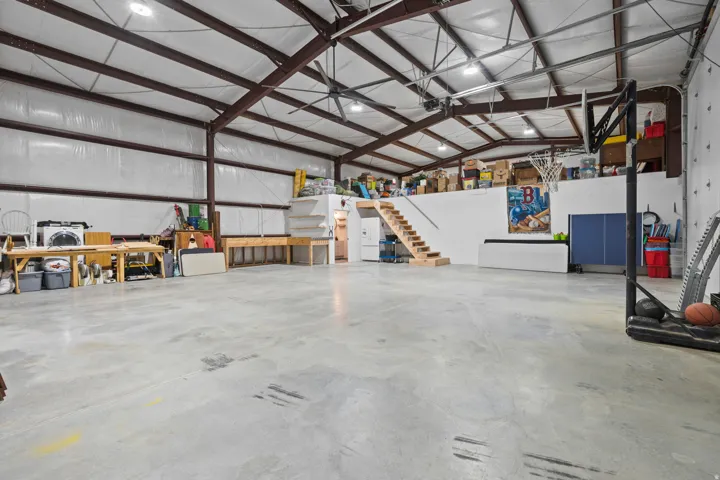 Spacious concrete-floored interior featuring high ceilings with exposed steel beams, industrial lighting, and a prominent mezzanine level with wood stairs