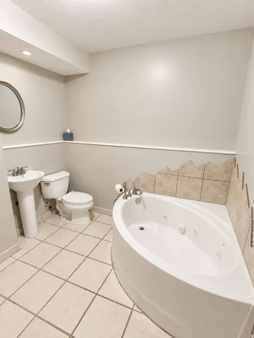 Corner jetted tub with tile surround, pedestal sink, toilet, and tile flooring