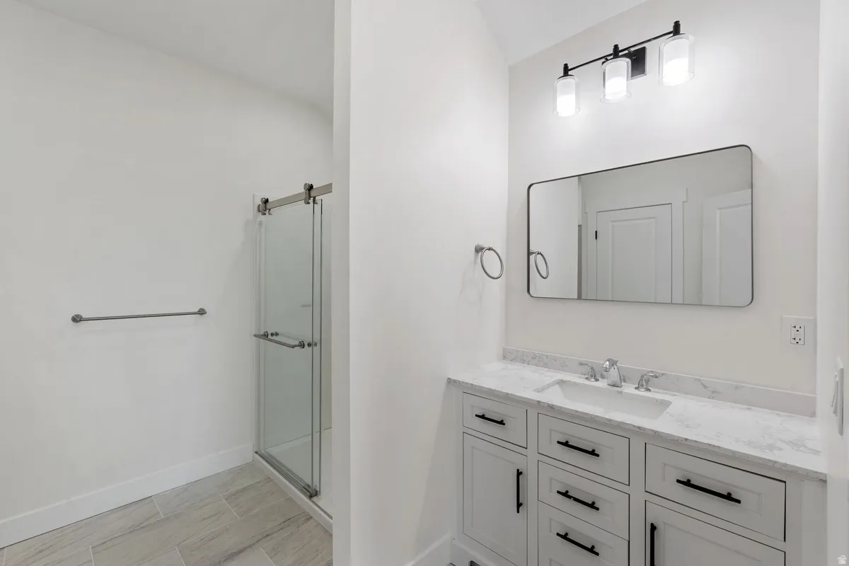 Bathroom featuring a glass-enclosed shower with a sliding barn door-style track, a single vanity with a white countertop and integrated sink, white cabinetry with black hardware, a rectangular mirror, and a three-light vanity fixture