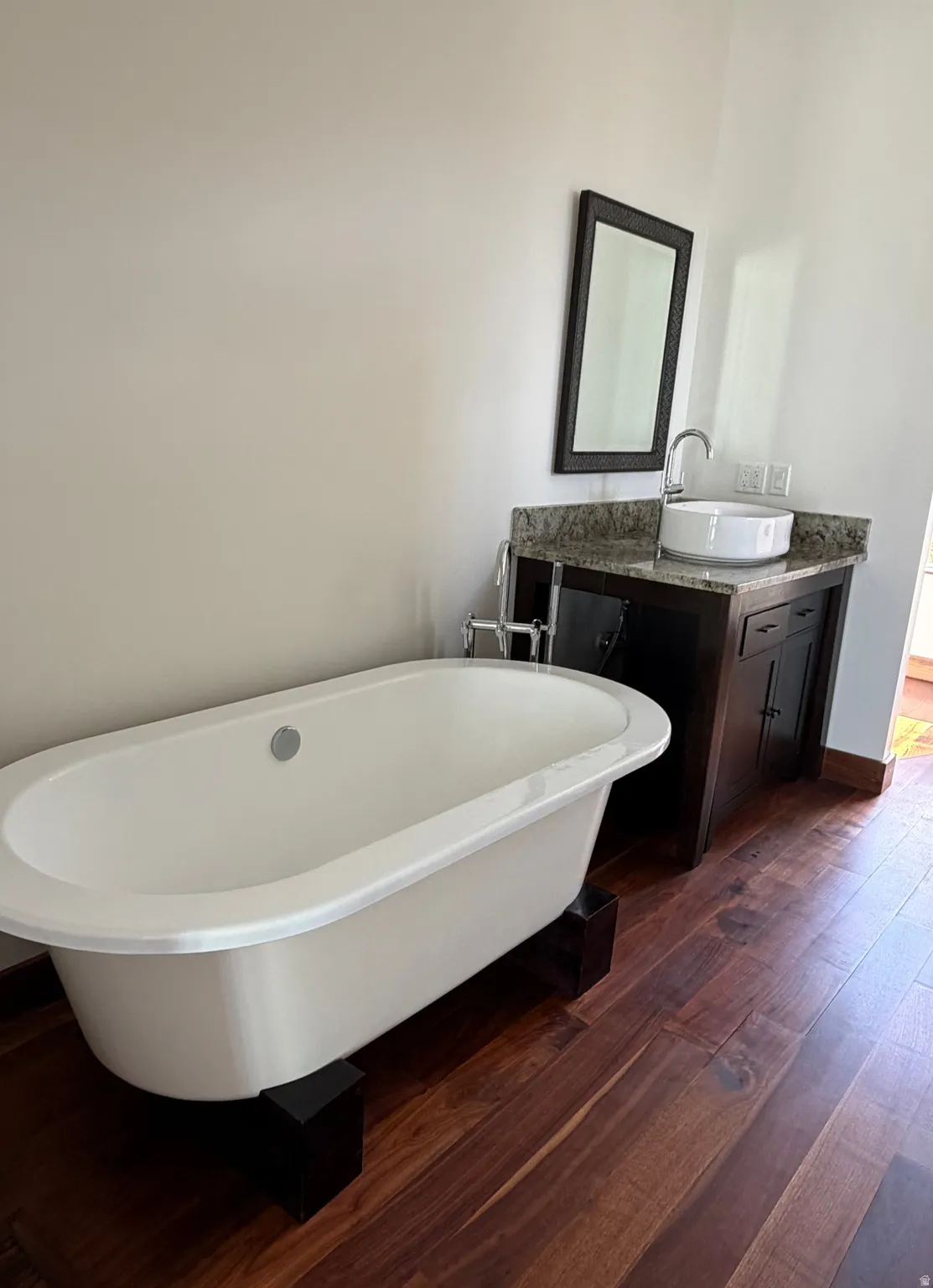 Freestanding soaking tub, wood-finish flooring, dark wood vanity with a stone countertop, and a vessel sink