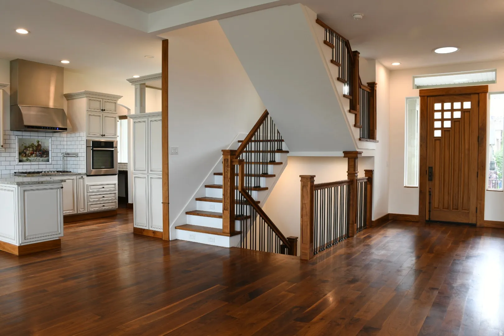 Hardwood flooring throughout, custom wood entry door, open-tread staircase with wrought iron balusters, recessed lighting, and kitchen with white cabinetry and stainless steel appliances
