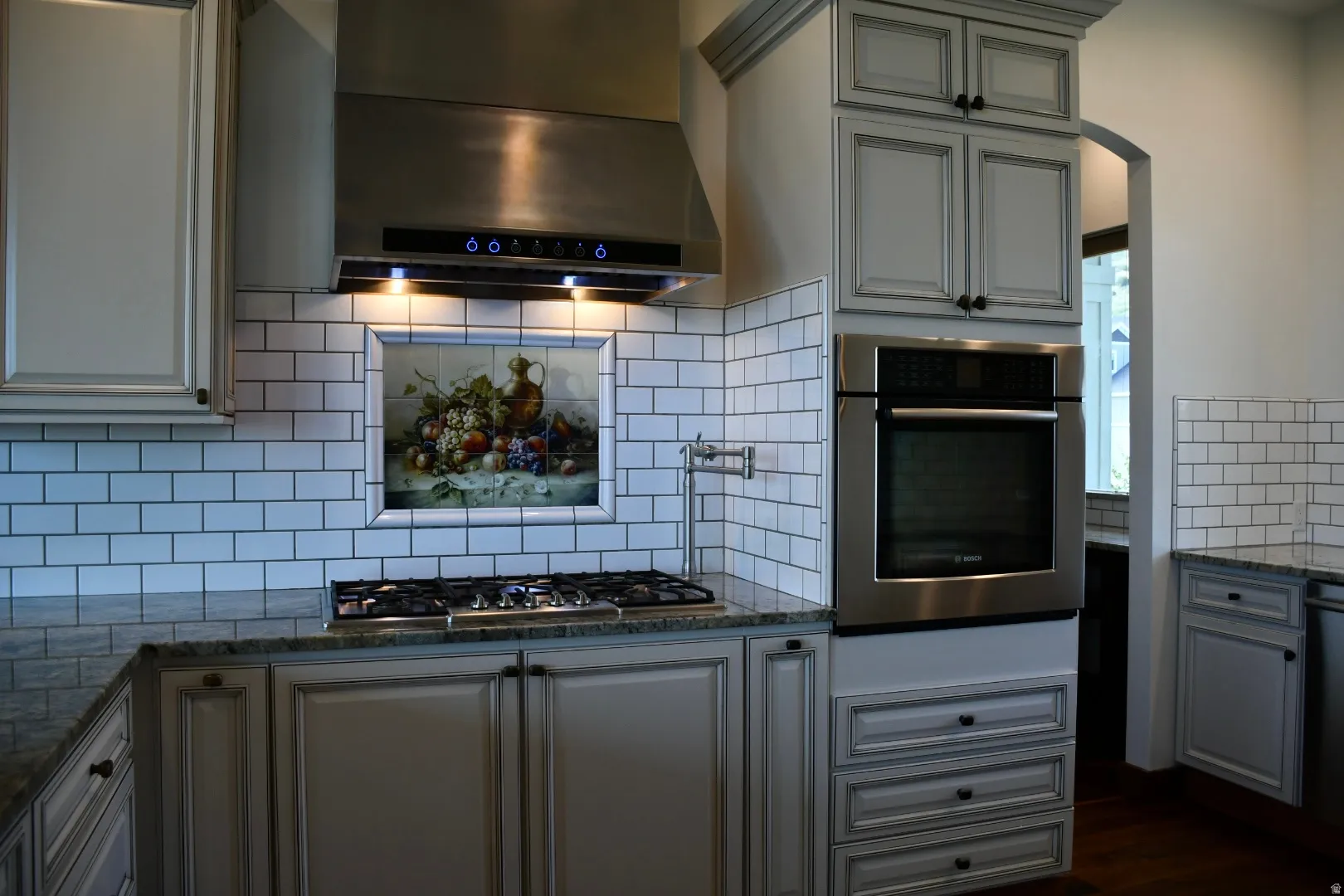 Stainless steel range hood, built-in wall oven, gas cooktop, subway tile backsplash, and granite countertops