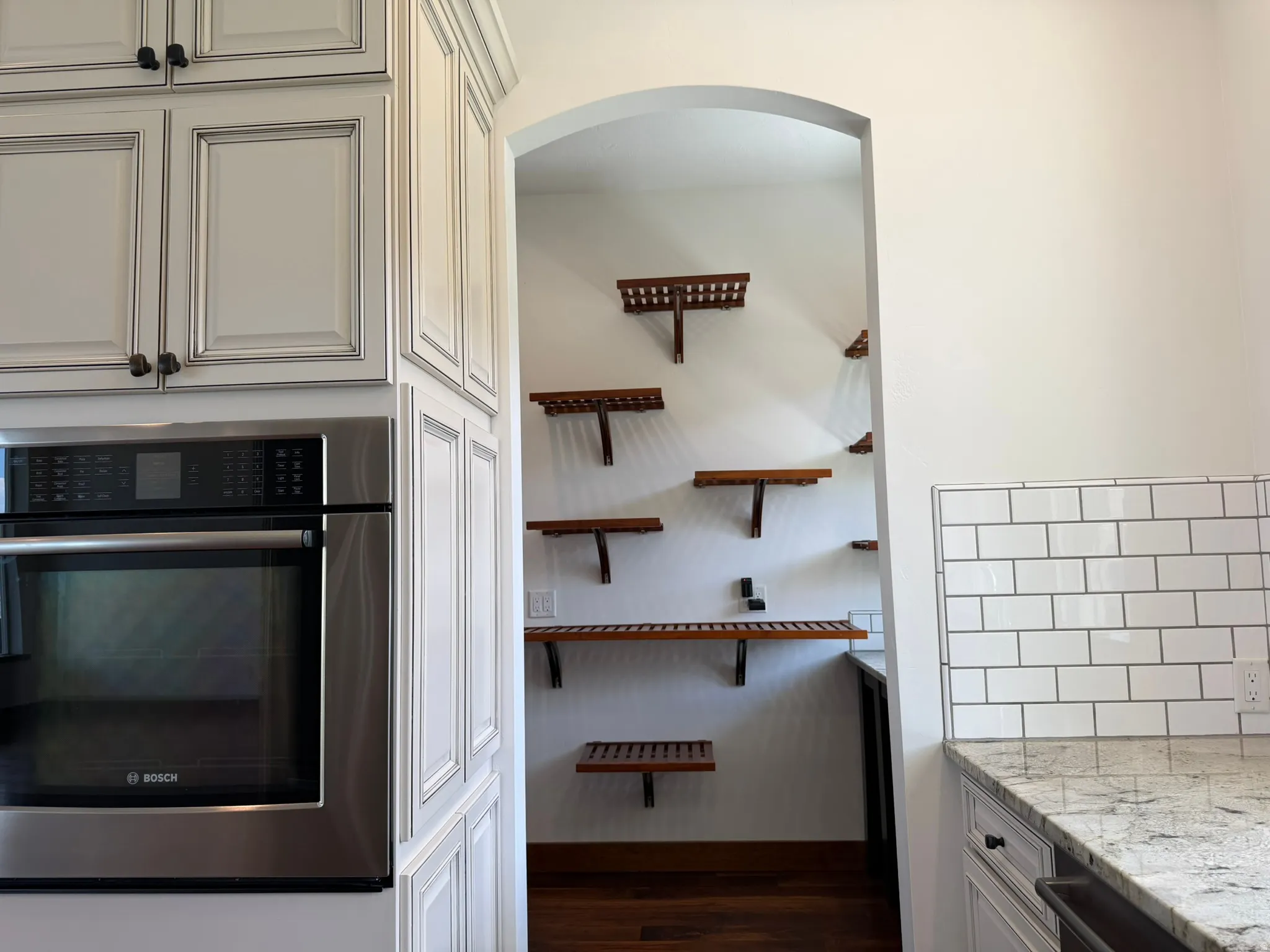 White paneled cabinetry, stainless steel built-in oven, granite countertop, white subway tile backsplash, and an arched doorway