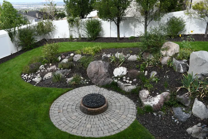 Manicured lawn, circular paver patio with fire pit, rock garden landscaping, mature shrubbery, and white privacy fencing