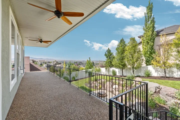 Spacious covered patio featuring exposed aggregate flooring, two ceiling fans, and black metal railings overlooking a landscaped yard with a fire pit area with an amazing view of the salt lake valley