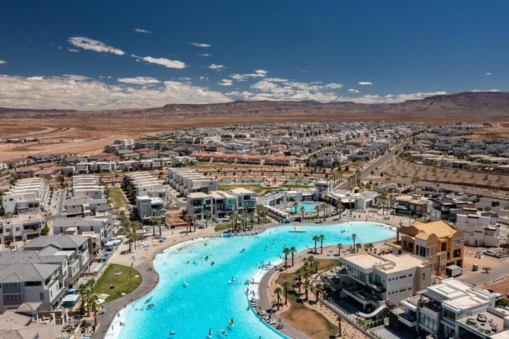 Expansive community featuring a large, blue water lagoon with surrounding sandy areas, numerous residential buildings, and distant mountain backdrops