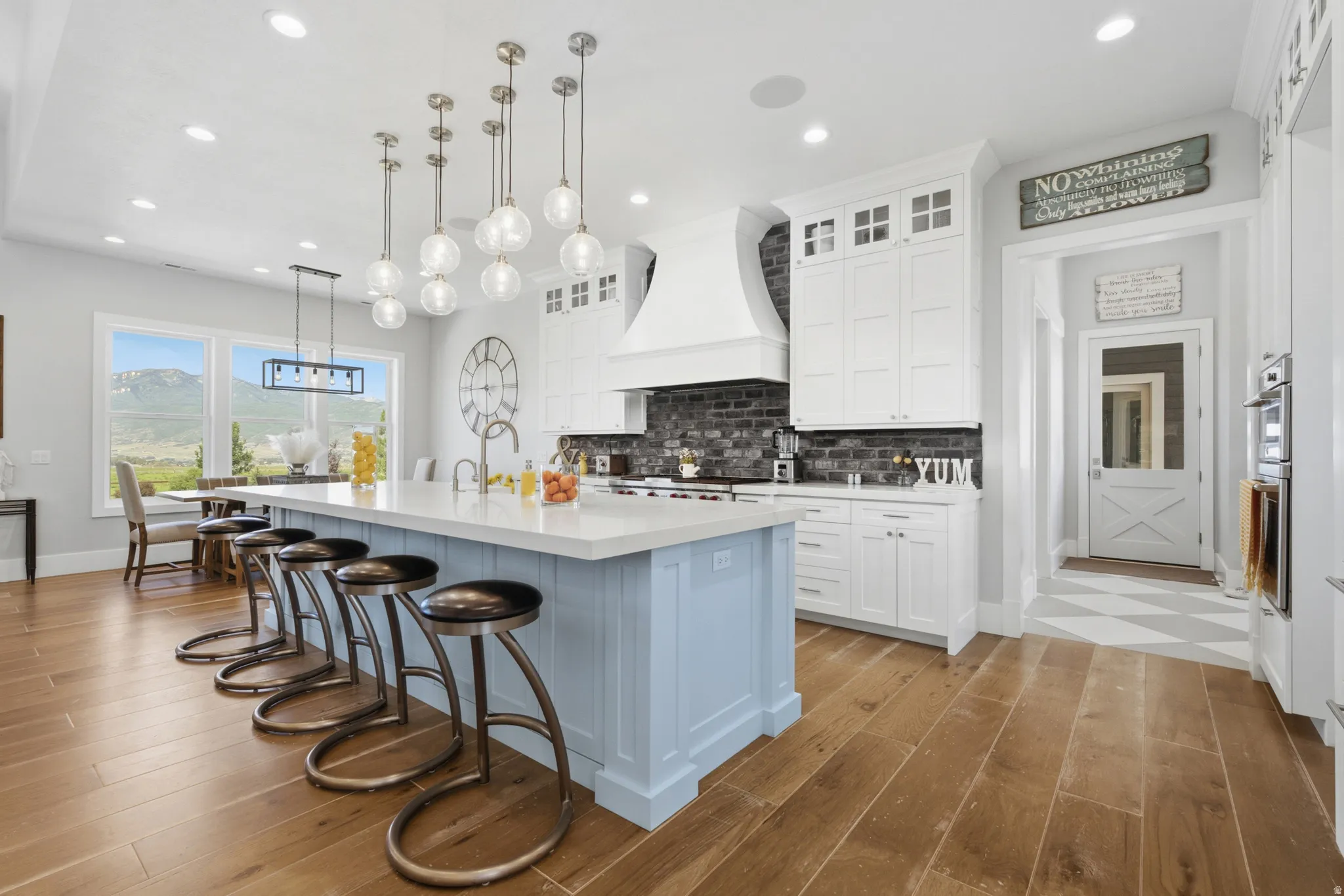 Spacious kitchen featuring a large island with seating, pendant lighting, wood-finish flooring, white cabinetry, and a dark brick-patterned backsplash